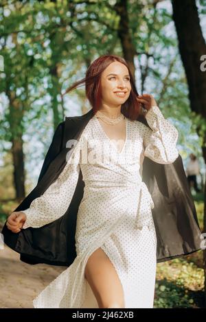 Hübsches rothaariges Mädchen in einem weißen Kleid und einer schwarzen Jacke, das an einem sonnigen Tag im Park spazieren geht Stockfoto