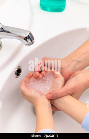 Seifenschaum in Herzform in den Kinderhände über dem Waschbecken und in den Händen der Mutter in einem hellen Badezimmer. Selektiver Fokus. Nahaufnahme Stockfoto