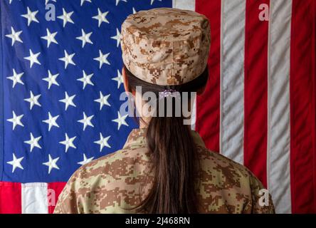 Soldatin der US-Armee, die vor einer amerikanischen Flagge steht. Frau in Militäruniform Rückansicht Stockfoto