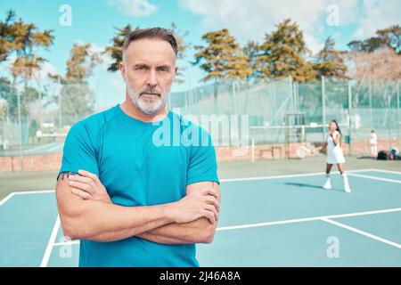 Ich Stelle sicher, dass mein Team sein Bestes gibt. Aufnahme eines hübschen, reifen Trainers, der während des Trainings mit gekreuzten Armen auf einem Tennisplatz steht. Stockfoto