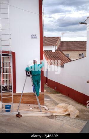 Ein Maler und Dekorateure malt ein Haus im Zentrum von Berrocal, Huelva, Spanien. Er trägt grüne Overalls und steht auf einer Leiter Stockfoto