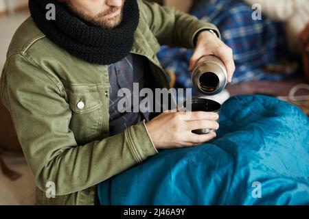 Nahaufnahme des Flüchtlings, der Tee aus Thermoskannen gießt, während er sich in einem mit Schlafsack bedeckten Unterschlupf versteckt und versucht, sich warm zu halten, Platz zum Kopieren Stockfoto