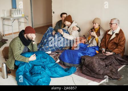 Gruppe von notleidenden kaukasischen Flüchtlingen, die sich während des Krieges oder der Krise in Unterschlupf versteckten, bedeckt mit Decken auf dem Boden Stockfoto