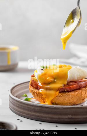 Gießen Hollandaise Sauce in Ei benedict mit pochiertem Ei, Schinken, Zwiebel auf Scheibe Toast für leckeres Frühstück. Vertikale Ausrichtung. Englischer Brunch Stockfoto