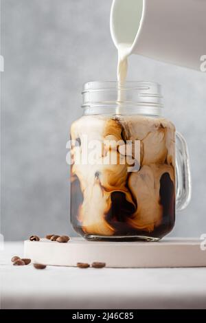 Gieße Milch oder Rahm in einem Glas auf grauem Hintergrund in den Eiskaffee. Kalte Erfrischung Sommergetränk. Vertikale Ausrichtung Stockfoto