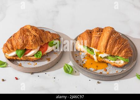 Croissant-Sandwiches mit pochiertem Ei, Avocado, Weichkäse, Mozzarella und Tomate auf einem Teller auf weißem Hintergrund. Französisches Frühstück. Leckeres Essen Stockfoto