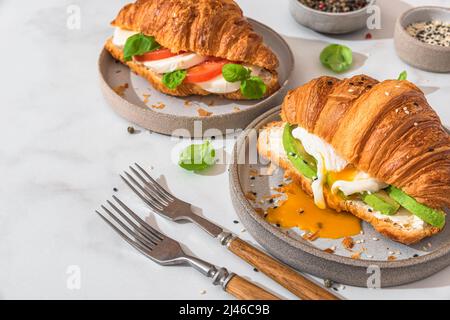 Leckeres Frühstück mit Croissant-Sandwiches mit pochiertem Ei, Avocado, Weichkäse, Mozzarella und Tomaten auf einem Teller mit Gabeln auf weißem Hintergrund Stockfoto