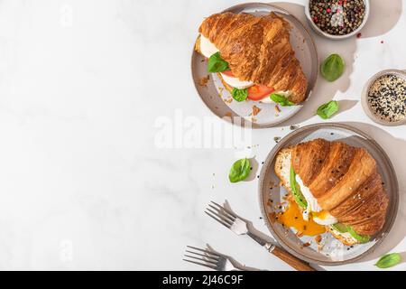 Croissant-Sandwiches mit pochiertem Ei, Avocado, Weichkäse, Mozzarella und Tomate auf einem Teller mit Gabeln auf weißem Hintergrund. Leckeres Frühstück. Oben Stockfoto