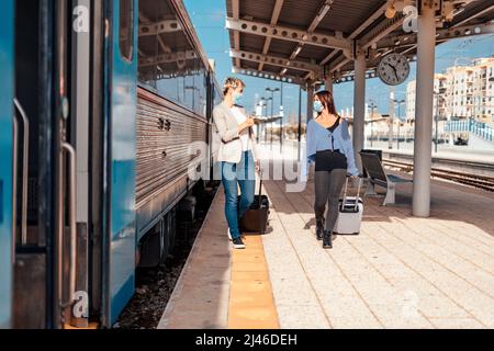 Zwei glückliche Freundinnen in Schutzmasken gehen und plaudern auf dem Bahnsteig am Bahnhof Stockfoto