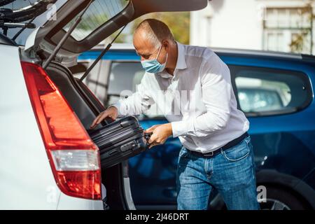 Ein Taxi- oder Uber-Fahrer in Maske legt das Gepäck in den Kofferraum seines Autos Stockfoto