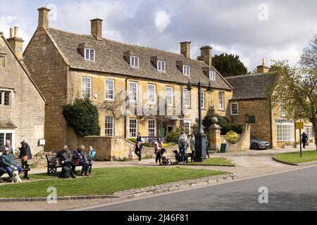 England Village - Straßenszene; Menschen, die die Frühlingssonne auf der High Street, Broadway Village, den Cotswolds, Worcestershire UK genießen Stockfoto