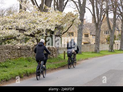 Zwei Menschen radeln durch das Dorf Cotswolds am Broadway und folgen im Frühjahr dem Evesham Blossom Trail, Broadway, Cotswolds Worcestershire UK Stockfoto