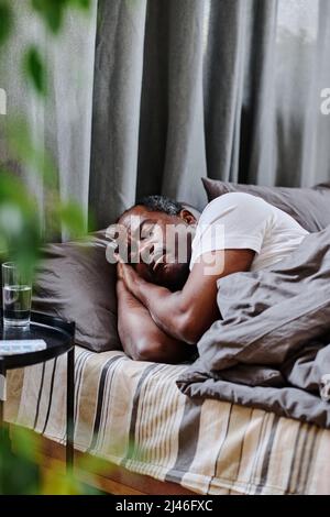 Müde reifer afroamerikanischer Mann, der morgens friedlich im Bett vor einem kleinen schwarzen Tisch mit einem Glas Wasser schlief Stockfoto