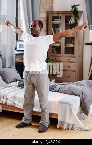 Aktiver afroamerikanischer Rentner in T-Shirt und Hose, der morgens mit ausgestreckten Armen Sport macht, während er am Bett steht Stockfoto