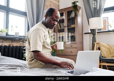 Ernsthafter pensionierter afroamerikanischer Mann mit einer Tasse Tee oder Kaffee, der auf den Laptop-Bildschirm schaut, während er auf dem Bett sitzt und Online-Nachrichten liest Stockfoto
