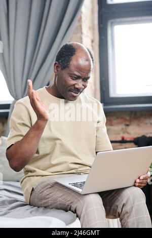 Ein glücklicher, älterer schwarzer Mann zu Hause trägt seiner Familie winkende Hand auf dem Laptop-Bildschirm, während er während der Online-Kommunikation auf dem Bett sitzt Stockfoto