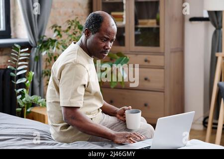 Seriöser afroamerikanischer Rentner, der während der Online-Kommunikation mit seiner Familie auf den Laptop-Bildschirm schaut, während er auf dem Bett sitzt Stockfoto