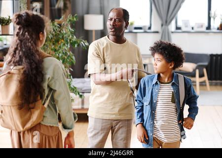 Älterer pensionierter afroamerikanischer Mann, der seinem Enkel beim Ausziehen des Rucksacks half, während er mit der Enkelin in einer Loft-Wohnung sprach Stockfoto