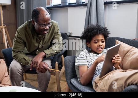 Glücklicher älterer afroamerikanischer Mann, der Online-Daten diskutiert, mit seinem niedlichen Enkel, der auf den Tablet-Bildschirm zeigt, während er im Sessel sitzt Stockfoto