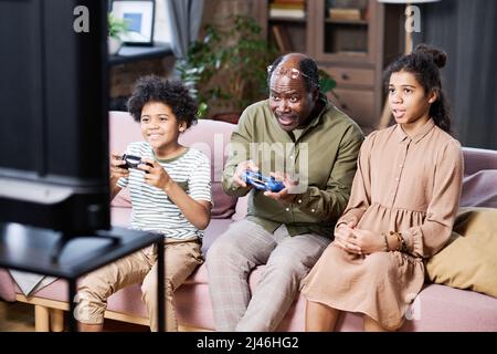 Eine Gruppe glücklicher Enkelkinder und ihr Großvater spielen vor dem fernseher, während sie auf der Couch im Wohnzimmer sitzen Stockfoto