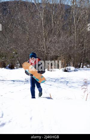 Fröhlicher Teenager-Mann, der auf dem Schnee läuft, während er ein Surfbrett hält Stockfoto