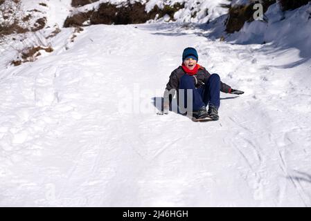 Junge rutscht vom Schneehang herunter. Genießen Sie die Winterrodelzeit Stockfoto