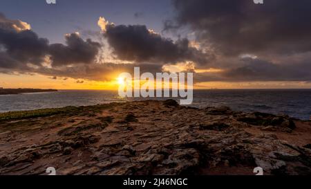 Felsige Küste bei Sonnenuntergang mit dramatischen Wolken und goldenem Licht über dem Meer. Raue Texturen und ein leuchtender Horizont schaffen eine malerische, stimmungsvolle Küste Stockfoto