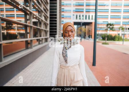 Selbstbewusste afroamerikanische Frau in modernem Outfit und muslimischer Hijab, die sich gegen moderne Wolkenkratzer im Business Center auflehnen und die Kamera betrachten Stockfoto