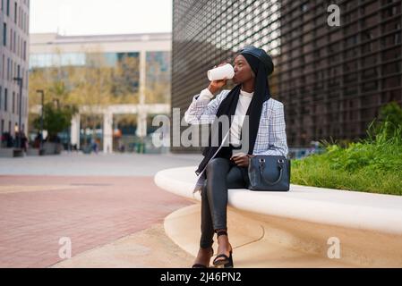 Stilvolle junge afroamerikanische Frau in elegantem legerem Outfit und schwarzem muslimischen Kopftuch, das auf einer Steinbank sitzt und Kaffee aus der Tasse im modernen Business Center trinkt Stockfoto