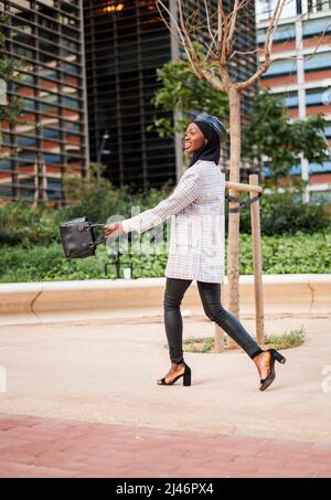 Seitenansicht Ganzkörper von sorglosen afroamerikanischen Frauen in trendigen formellen Outfit und muslimischen Kopftuch genießen Spaziergang im Stadtpark und lächeln glücklich Stockfoto