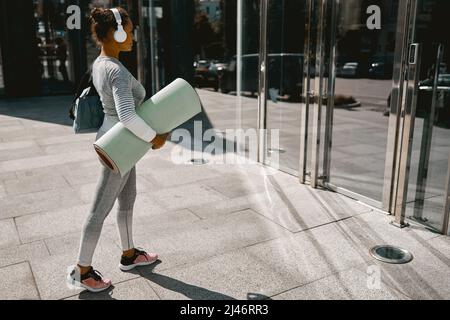 Sportliche Yoga-Frau in aktiver Kleidung und Kopfhörer beim Gehen mit Matte für das Training Stockfoto