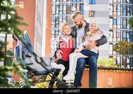 Fröhliche Familie umarmt sich, verbringt Zeit im Freien, während das Baby im Kinderwagen schläft. Glücklicher Vater sitzt auf der Bank mit Sohn und Tochter im modernen Innenhof der Stadt Wohnhochhäuser. Stockfoto