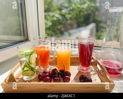 Holztablett mit bunten Getränken, Kirschen auf einer Fensterbank. Dekanter mit den Resten von rotem Saft. Offenes Fenster, heißer Sommer, Sonnenlicht. Stockfoto