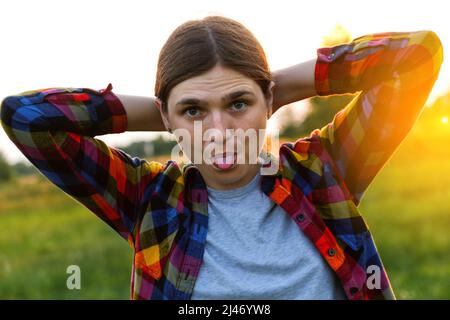 Unschärfe lustige junge Frau zeigt Zunge und Blick auf die Kamera auf Natur Sonnenuntergang Hintergrund draußen. Nahaufnahme Porträt Mädchen Ausdruck, Emotion. Aus Stockfoto