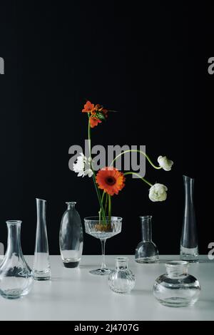 Vertikale zeitgenössische Stillleben Komposition aus bunten Blumen und verschiedenen transparenten Glasgefäßen auf grauer Tischfläche vor schwarzem Wandhintergrund Stockfoto