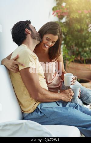 Flüsternde Liebe in ihr Ohr. Aufnahme eines glücklichen jungen Paares, das auf einer Bank draußen Kaffee trinken kann. Stockfoto