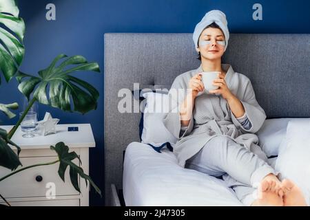 Frau in Pyjama und Bademantel mit Augenflecken und Handtuch auf ihrem Kopf, entspannen und trinken Morgenkaffee, während sie auf ihrem Bett sitzt. Morgendliche Gewohnheiten und Stockfoto