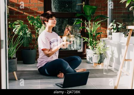 Junge Frau, die Yoga-Übungen macht und auf einer tibetischen Klangschale mit Schlägel in einem gemütlichen, modernen Loft-Zimmer voller heimeliger Pflanzen am sonnigen Morgen spielt. Stockfoto