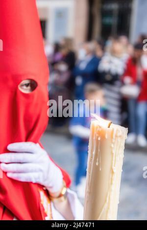 Die brennende Kerze eines jungen Nazareners mit einer roten Kapuze, der in der Karwoche seine Buße tat. Selektiver Fokus nur mit Kerzenflamme Stockfoto