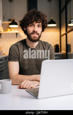 Vertikales Porträt eines glücklichen bärtigen jungen Geschäftsmanns, der auf dem Laptop am Tisch in einem modernen Küchenraum sitzt und die Kamera anschaut. Stockfoto