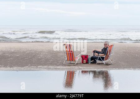 Reifer Mann mit grauen Haaren sitzt auf einem Strandstuhl lächelnd, während er die Kamera anschaut. Das Meer im Hintergrund. Stockfoto
