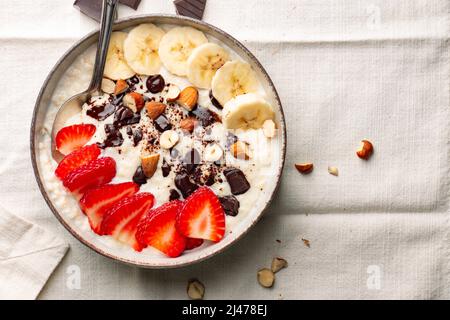 Hirsebrei mit Erdbeere, Banane, dunklen Schokoladenstücken, Haselnuss und Mandel in einer Keramikschale auf einer weißen Serviette Stockfoto