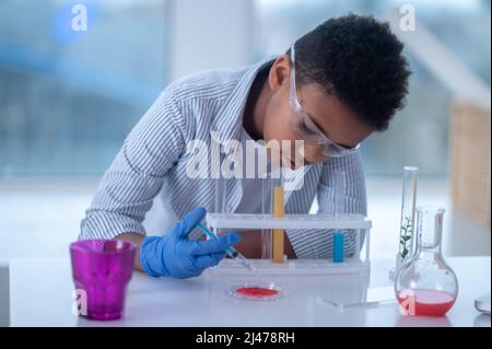 Ein dunkelhaariger Junge in einem Labormantel, der eine Spritze mit einem Reagenz hält und beschäftigt aussieht Stockfoto