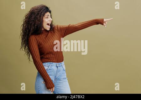 SAISONALES SONDERANGEBOT. Fröhliche, lächelnde lockige lateinische Frau bleibt nahe der grünen Wand, sagt Hey, schau dir das an, schau zur Seite, zeige an freiem Platz den Kopierraum Stockfoto