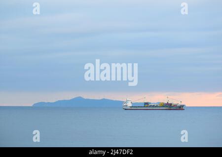 Frachtschiff vor einer Insel (mit Luftturbinen) bei Sonnenuntergang. Ägäis und blaue Wolken Stockfoto