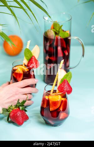 Weibliche Hand hält ein Glas mit traditionellem spanischen Sangria-Cocktail, verziert mit Stücken frischer Erdbeere, einer Prise Minze und Stroh aus Öko-Papier. Stockfoto