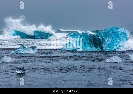 Ozeanwellen brechen über Eisberge und große Stücke blauen Eises an einem vulkanischen schwarzen Sandstrand (Diamond Beach, Island) Stockfoto