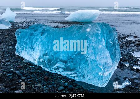 Ozeanwellen brechen über Eisberge und große Stücke blauen Eises an einem vulkanischen schwarzen Sandstrand (Diamond Beach, Island) Stockfoto