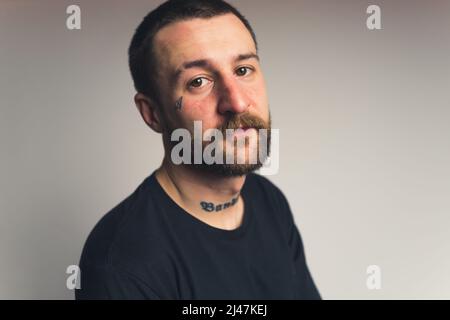 Hübscher bärtiger tätowierte europäische junge Mann mit einem Schnurrbart, der ernsthaft mittel Nahaufnahme Gradient Hintergrund Studioaufnahme aussieht. Hochwertige Fotos Stockfoto