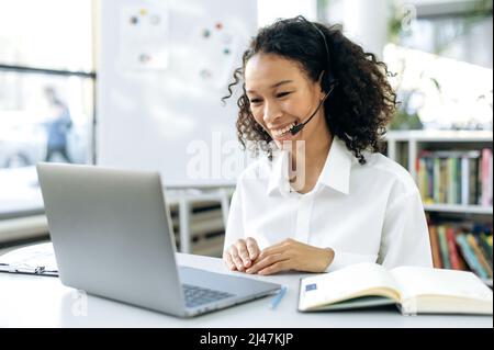 Positive, intelligente afroamerikanische Frau mit Headset, Supportmitarbeiter, Online-Berater, sitzen am Desktop vor einem Laptop, in einem modernen Büro, mit einem Videoanruf mit dem Kunden, lächelnd freundlich Stockfoto